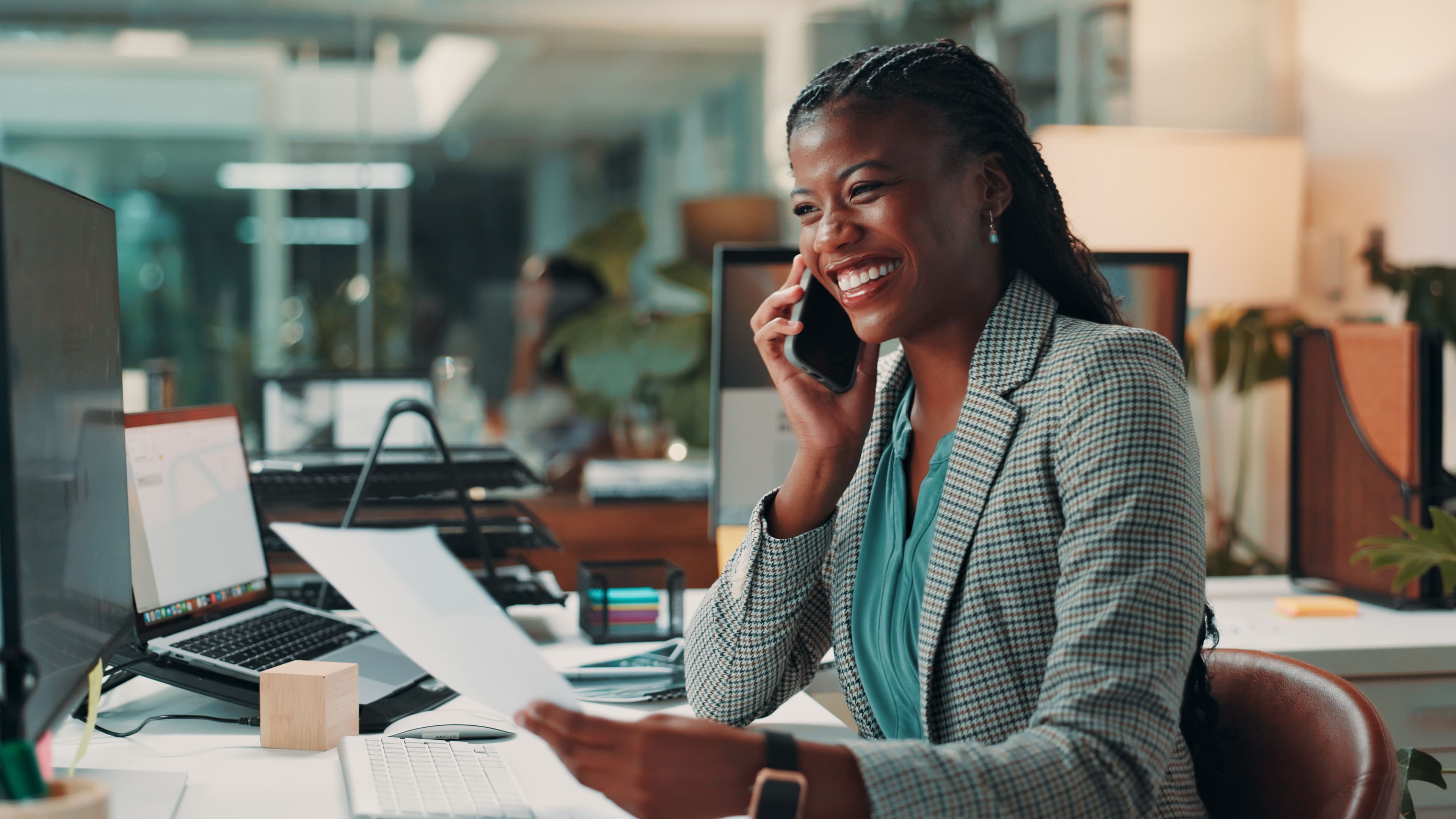 business woman sat at her desk on the phone whilst smiling and holding a piece of paper
